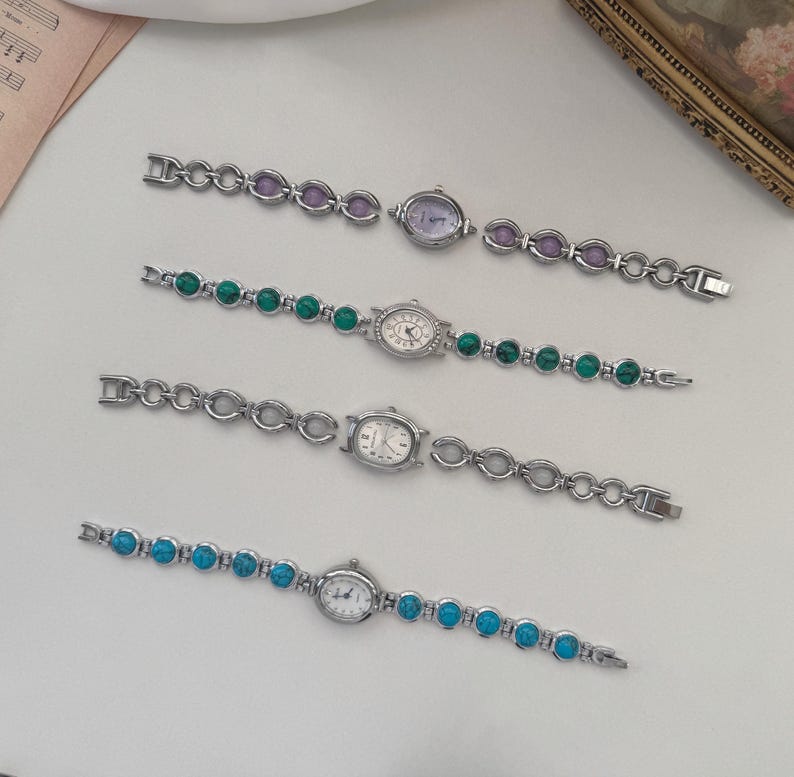 May include: Four silver-toned wristwatches with different colored stone accents. The watches have oval or round faces and matching stone bracelets in purple, green, and turquoise. The watches are displayed on a white surface.