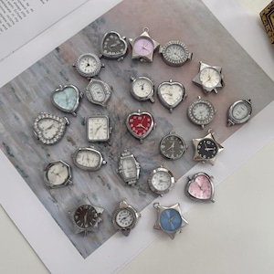 Puede incluir: Una colección de pequeñas esferas de reloj decorativas en varias formas, incluyendo corazones, estrellas y círculos. Las esferas tienen marcos de metal plateado y diferentes colores, incluyendo blanco, negro y rosa. Algunas tienen detalles decorativos.