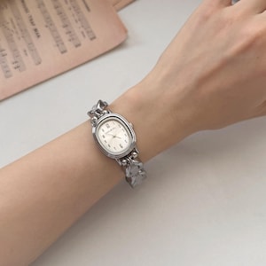 May include: A silver-toned wristwatch with an oval face and a linked band. The watch face is white with black numerals and hands. The band has a textured, organic design. The watch is on a wrist.