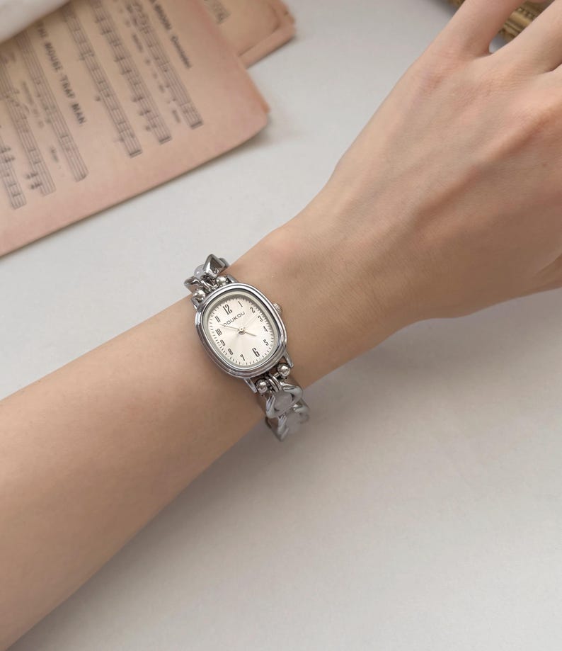 May include: A silver-toned wristwatch with an oval face and a linked band. The watch face is white with black numerals and hands. The band has a textured, organic design. The watch is on a wrist.