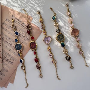 May include: Five gold-toned, decorative wristwatches with various colored gemstones and watch faces. The watches have different shaped faces, including round, square, and heart-shaped. The watch bands are adorned with colorful beads and charms.