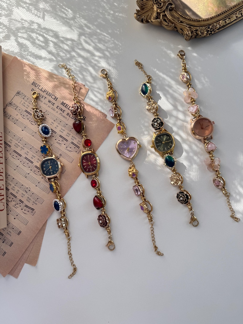May include: Five gold-toned, decorative wristwatches with various colored gemstones and watch faces. The watches have different shaped faces, including round, square, and heart-shaped. The watch bands are adorned with colorful beads and charms.