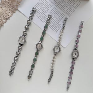 Puede incluir: Cuatro elegantes relojes de mujer con correas de metal plateado. Las esferas de los relojes son ovaladas o redondas, con perlas, piedras verdes y detalles de piedras rosas. Los relojes se exhiben sobre una superficie blanca.
