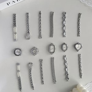 May include: A collection of silver-coloured watch components and watch faces are arranged on a white surface. The collection includes watch straps with pearl accents, chain links, and various watch faces in round, oval, and heart shapes.