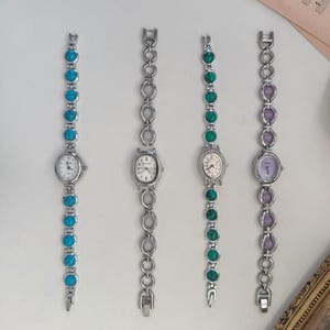May include: Four silver-toned wristwatches with oval or round faces and matching link bracelets. The bracelets feature turquoise, green, and purple stones. The watch faces have white dials and black numerals or markers. The watches are displayed on a white surface.