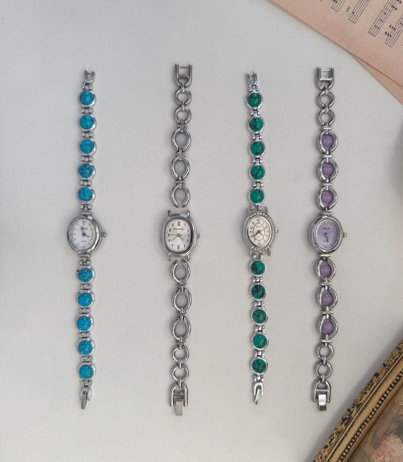May include: Four silver-toned wristwatches with oval or round faces and matching link bracelets. The bracelets feature turquoise, green, and purple stones. The watch faces have white dials and black numerals or markers. The watches are displayed on a white surface.