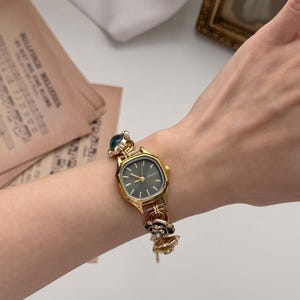 May include: Gold-toned wristwatch with a dark green face and decorative floral and jewel accents. The watch has a rectangular face with a gold bezel and a delicate chain-link band. The watch is worn on a wrist.