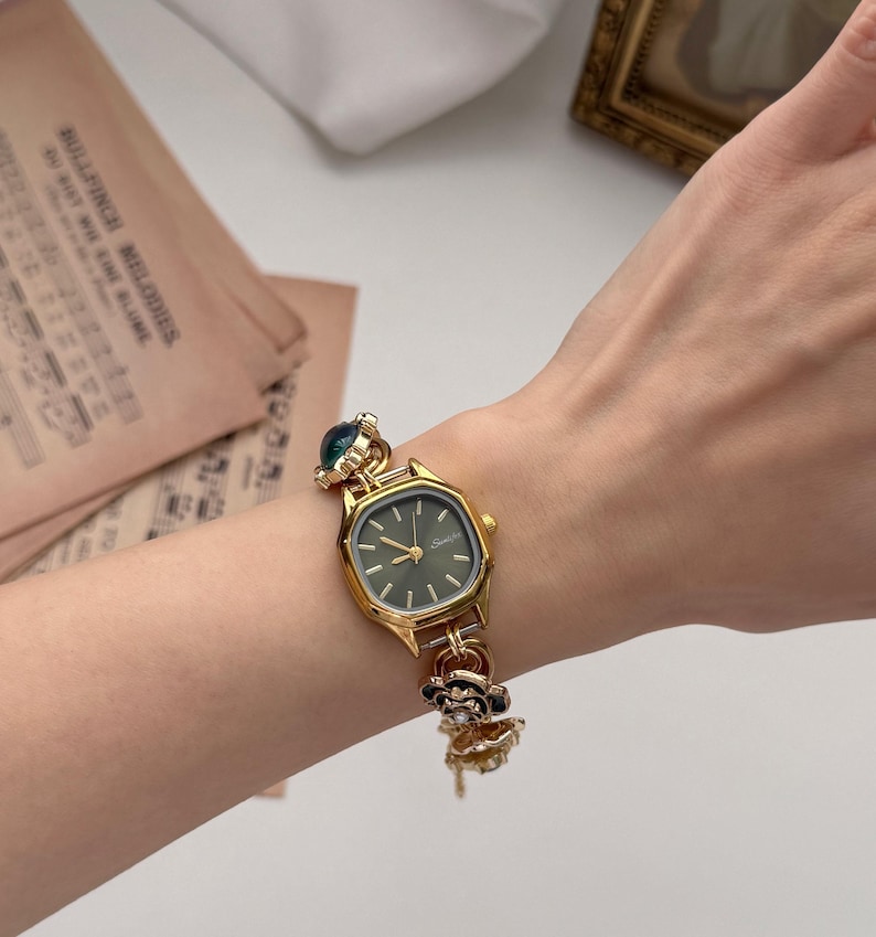 May include: Gold-toned wristwatch with a dark green face and decorative floral and jewel accents. The watch has a rectangular face with a gold bezel and a delicate chain-link band. The watch is worn on a wrist.