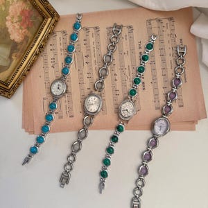 May include: Four vintage-style wristwatches with oval faces and silver-tone metal bands. Each watch features a different gemstone: turquoise, clear, green, and purple. The watches rest on a sheet of aged sheet music.