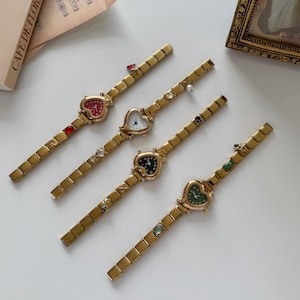 Puede incluir: Cuatro relojes dorados en forma de corazón con correas doradas a juego. Cada esfera del reloj tiene un color diferente: rojo, blanco, negro y verde. Las correas están adornadas con varias piedras preciosas. Estética vintage.