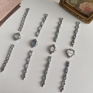 May include: A collection of silver-toned wristwatches and matching bracelet segments arranged on a surface. The set includes various watch faces, including round, oval, and heart-shaped designs, paired with link bracelets featuring iridescent stone inlays.