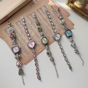 Puede incluir: Cinco pulseras de plata con dijes y diferentes esferas de reloj, incluyendo formas de corazón y cuadradas. Las pulseras presentan detalles en rosa, verde y turquesa. El fondo incluye partituras y un libro.