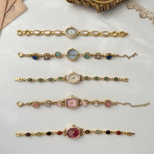 Op de afbeelding: Vijf goudkleurige polshorloges horizontaal gerangschikt. Het bovenste horloge heeft witte ovale schakels. De tweede heeft blauwe en turquoise bloemaccenten. De derde heeft groene ovale stenen. De vierde toont roze hartvormige schakels. Het onderste horloge heeft rode en bruine stenen.