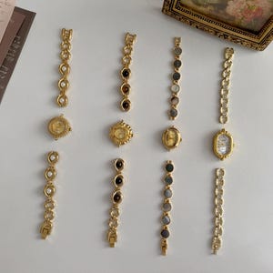 May include: An assortment of vintage gold-toned watch faces and watch bands. The watch faces have intricate designs and are paired with bands featuring pearls, gemstones, and chain links. The pieces are arranged on a white surface.