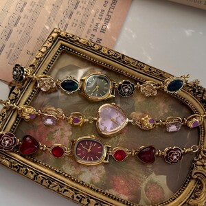 May include: Three vintage-style gold-toned watches with decorative bands. The watches have different colored faces, including green, pink, and red. The bands feature floral and gemstone accents. The watches are displayed in a gold-framed tray.