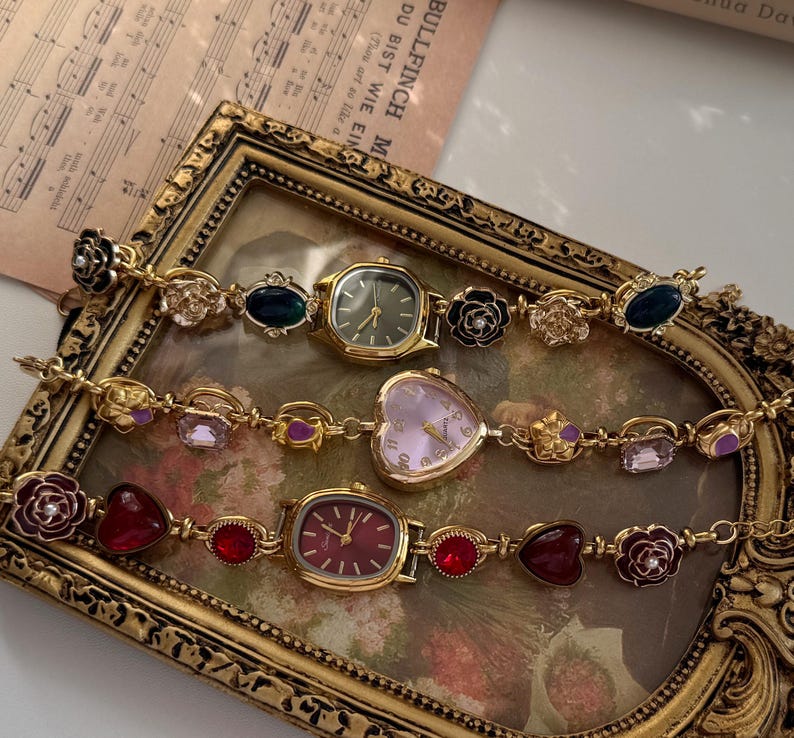 May include: Three vintage-style gold-toned watches with decorative bands. The watches have different colored faces, including green, pink, and red. The bands feature floral and gemstone accents. The watches are displayed in a gold-framed tray.