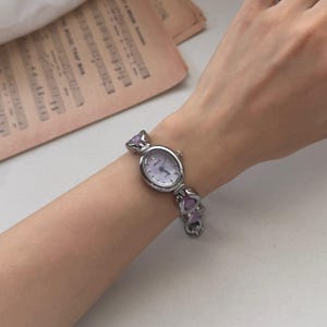 May include: A silver-toned wristwatch with a light purple face and matching purple accents. The watch has an oval face and a linked band with heart-shaped purple embellishments. The watch is on a wrist.