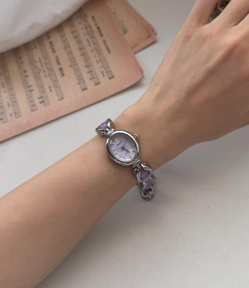 May include: A silver-toned wristwatch with a light purple face and matching purple accents. The watch has an oval face and a linked band with heart-shaped purple embellishments. The watch is on a wrist.