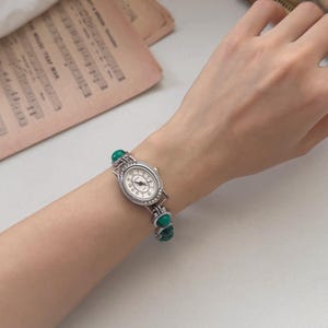 May include: Silver-toned wristwatch with an oval face and a white dial featuring black numerals. The watch is adorned with small, clear crystals around the face and has a band with green stone accents. The watch is on a wrist.