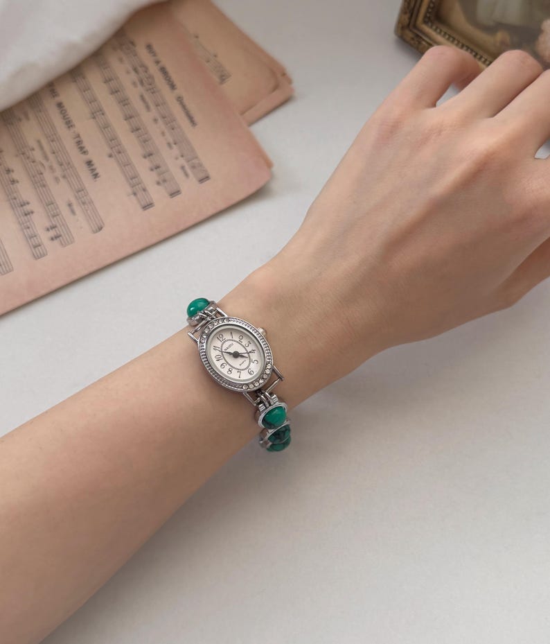 May include: Silver-toned wristwatch with an oval face and a white dial featuring black numerals. The watch is adorned with small, clear crystals around the face and has a band with green stone accents. The watch is on a wrist.