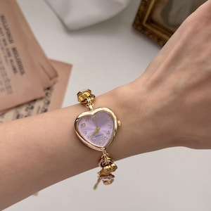 May include: A gold-toned quartz watch with a heart-shaped face and a light purple dial. The watch has a delicate chain and decorative charms. The watch is worn on a wrist.