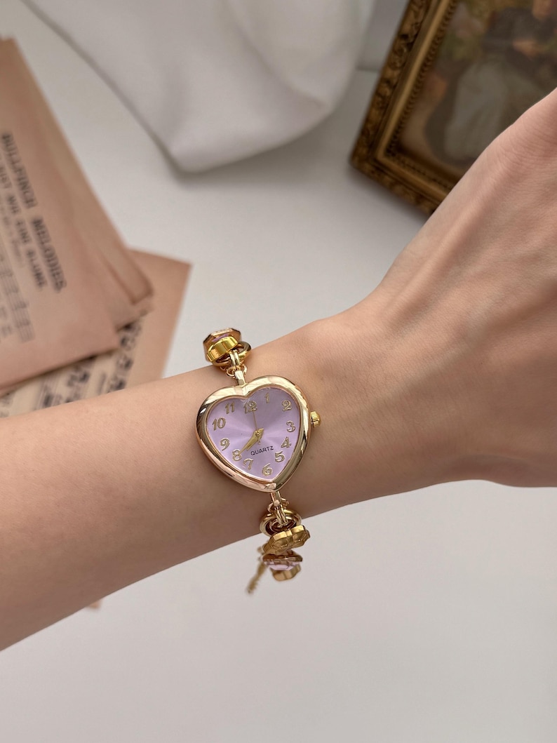 May include: A gold-toned quartz watch with a heart-shaped face and a light purple dial. The watch has a delicate chain and decorative charms. The watch is worn on a wrist.