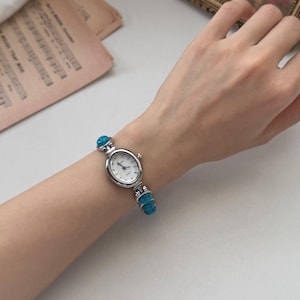 May include: A silver-toned wristwatch with a white face and turquoise accents. The watch has an oval face and a band adorned with turquoise stones. The watch is displayed on a person's wrist.