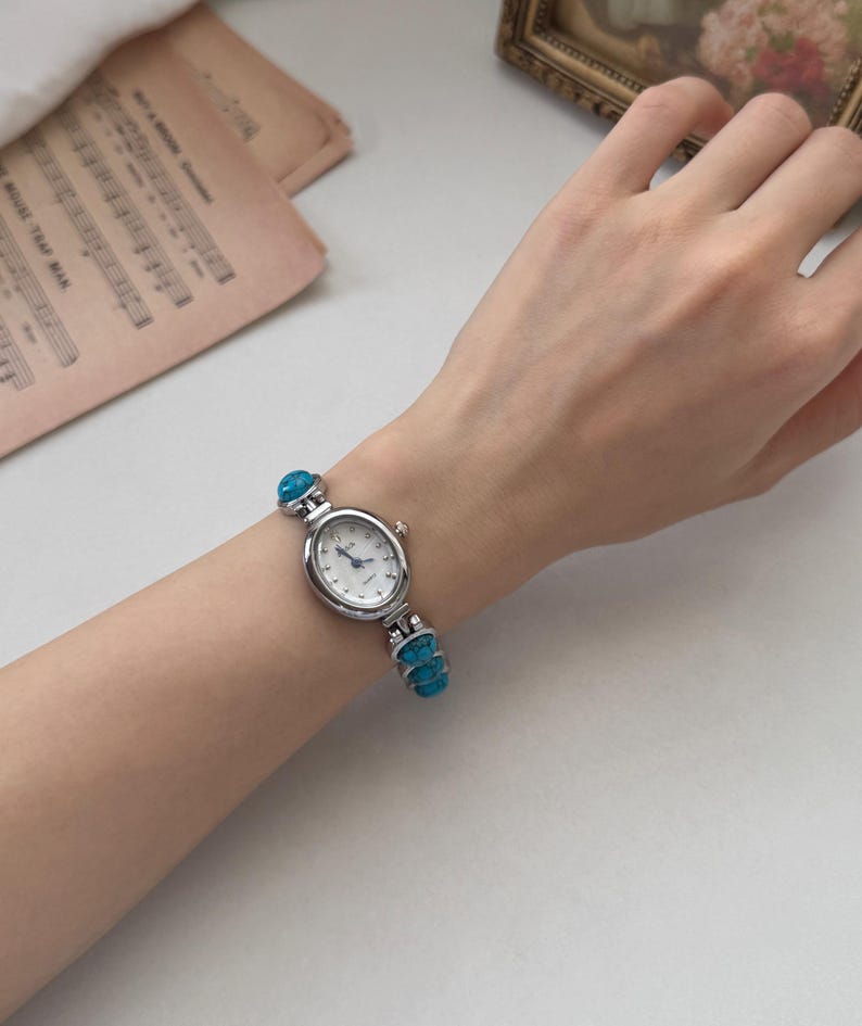 May include: A silver-toned wristwatch with a white face and turquoise accents. The watch has an oval face and a band adorned with turquoise stones. The watch is displayed on a person's wrist.