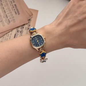 May include: A gold-toned wristwatch with a navy blue face and a decorative band featuring blue floral accents. The watch has a rectangular face with the text "MIGAI" and the numbers 1 through 12. The watch is on a wrist.