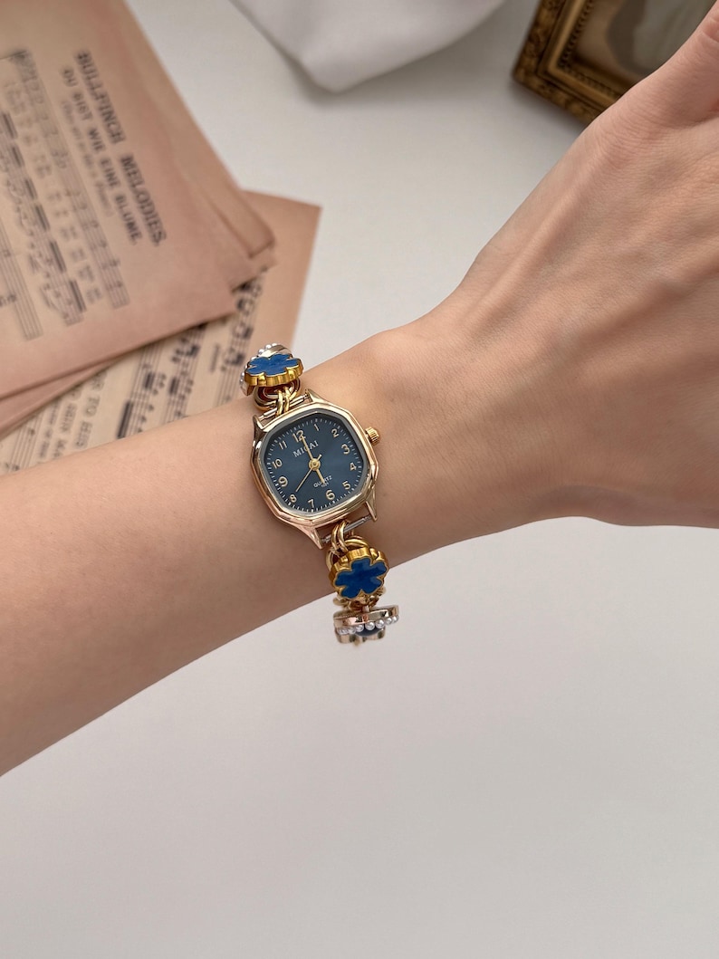 May include: A gold-toned wristwatch with a navy blue face and a decorative band featuring blue floral accents. The watch has a rectangular face with the text "MIGAI" and the numbers 1 through 12. The watch is on a wrist.