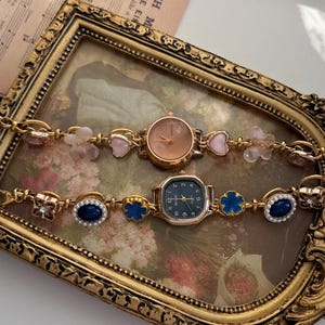May include: Two gold-toned wristwatches with decorative bands. One watch has a round, copper-colored face, while the other has a square, dark blue face. The bands feature pink hearts, blue flowers, and oval blue gems.