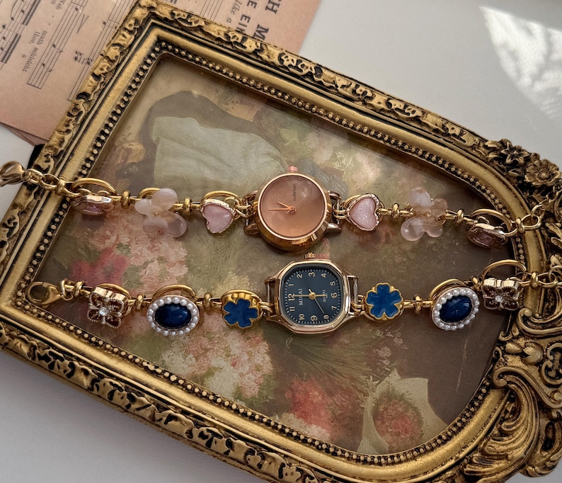 May include: Two gold-toned wristwatches with decorative bands. One watch has a round, copper-colored face, while the other has a square, dark blue face. The bands feature pink hearts, blue flowers, and oval blue gems.
