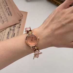 May include: A rose gold wristwatch with a round face and a delicate floral design. The watch features a light pink dial and is adorned with small, pink flower charms on the band. The watch is on a wrist.