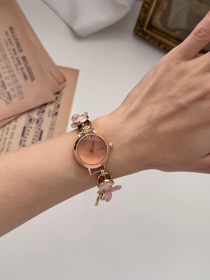 May include: A rose gold wristwatch with a round face and a delicate floral design. The watch features a light pink dial and is adorned with small, pink flower charms on the band. The watch is on a wrist.