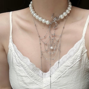 May include: A pearl necklace with a silver spider pendant and a dangling web design. The necklace features large, round pearls and a delicate, intricate web with small silver beads and chains. The spider is a stylized, skeletal design.