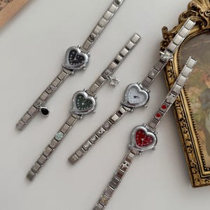 Op de afbeelding: Vier zilverkleurige horloges met hartvormige wijzerplaten en bijpassende schakelbanden. De wijzerplaten van de horloges zijn zwart, groen, wit en rood. Elk horloge heeft een ander ontwerp op de band.