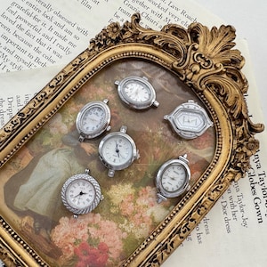 May include: A collection of small, silver-toned watch faces with white dials and various designs. The watches are displayed inside a decorative gold-colored frame, set against a backdrop of a vintage painting and text.