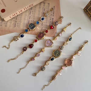Puede incluir: Cinco relojes de pulsera dorados con esferas de colores y abalorios decorativos. Los abalorios incluyen corazones, flores y piedras preciosas en tonos rojos, azules, rosas y verdes. Los relojes se exhiben sobre una superficie blanca.