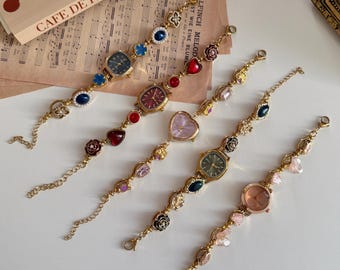 May include: Five gold-toned, decorative wristwatches with various colored watch faces and decorative charms. The charms include hearts, flowers, and gemstones in shades of red, blue, pink, and green. The watches are displayed on a white surface.