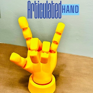 Może przedstawiać: Jasnożółta, przegubowa dłoń w geście rock and rolla. Dłoń jest drukowana w 3D i posiada ruchome stawy. Podstawa jest okrągła z czerwonym akcentem. Tekst "Articulated HAND" znajduje się nad dłonią w kolorze niebieskim i białym.