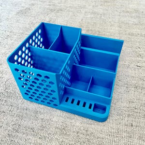 Puede incluir: Un organizador de escritorio azul brillante con múltiples compartimentos y un lado perforado. El organizador tiene secciones para bolígrafos, lápices y otros útiles de oficina. Está hecho de un material sólido y se encuentra sobre una alfombra gris claro.