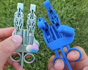 May include: Two articulated mechanical hand toys with finger loops at the base. One toy features a multi-coloured iridescent finish, while the other is solid blue. Both devices have segmented, jointed finger extensions that move independently.