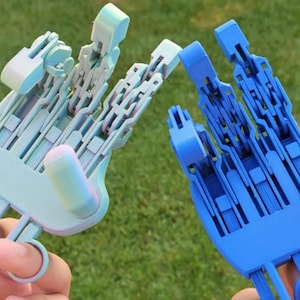 May include: Two 3D-printed prosthetic hands, one light blue and one dark blue, are held up against a green background. The hands are detailed with articulated fingers and a ring-like attachment for the user's fingers.