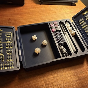 May include: A dark blue, tri-fold tabletop gaming organiser featuring integrated mechanical trackers for stats and skills. The left panel displays numerical dials for attributes, while the right panel lists skills. The centre holds three dice, a miniature figure, and pens.