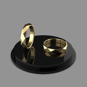 May include: Two gold-colored rings with geometric, faceted designs. One ring stands upright, while the other rests on a black, circular display stand. The rings reflect light, showcasing their intricate patterns. The background is a solid gray.