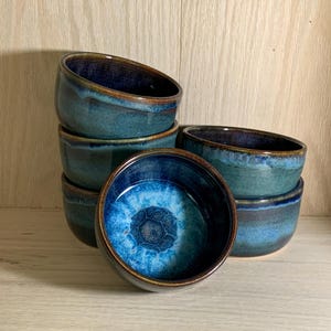 Puede incluir: Un conjunto de seis cuencos de cerámica hechos a mano. Los cuencos están esmaltados en tonos azules y turquesas, con un acabado brillante. El interior de un cuenco presenta un patrón texturizado único. Los cuencos están colocados sobre una superficie de madera clara.