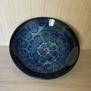 May include: A round ceramic bowl featuring a deep blue glaze with a reactive finish. The interior displays a distinct, repeating circular pattern of light blue and cream-colored petal-like shapes that radiate from the center toward the rim.