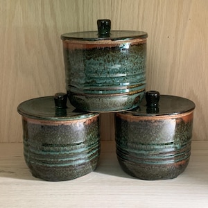 Puede incluir: Tres tarros de cerámica con tapas. Los tarros son cilíndricos con un esmalte verde oscuro y marrón. Cada tarro tiene una tapa a juego con un pequeño pomo negro redondo. Los tarros están apilados sobre una superficie de madera.