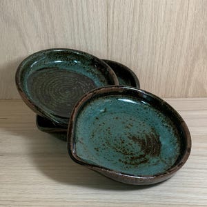 May include: A stack of four handmade ceramic spoon rests. The spoon rests have a unique, organic shape with a teal and brown glaze. The rims are a darker brown, and the interior has a speckled pattern. The background is a light wood.