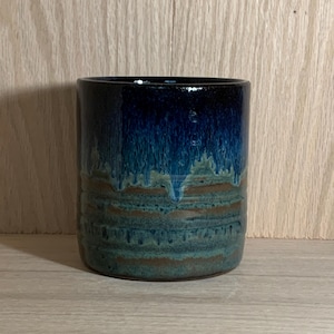 Może przedstawiać: Ręcznie robiony ceramiczny kubek z błyszczącym, warstwowym szkliwem. Góra jest w kolorze głębokiego granatu, który przechodzi w odcienie turkusu i brązu w kierunku podstawy. Cylindryczny kubek nadaje się do gorących lub zimnych napojów.